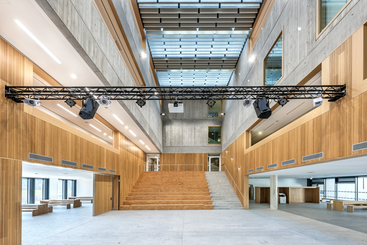 Zentraler Blick in die mehrstöckige Aula mit Obertageslicht, breiter Sitztreppe aus Holz und einer Gestaltung aus Sichtbeton und Holzwänden