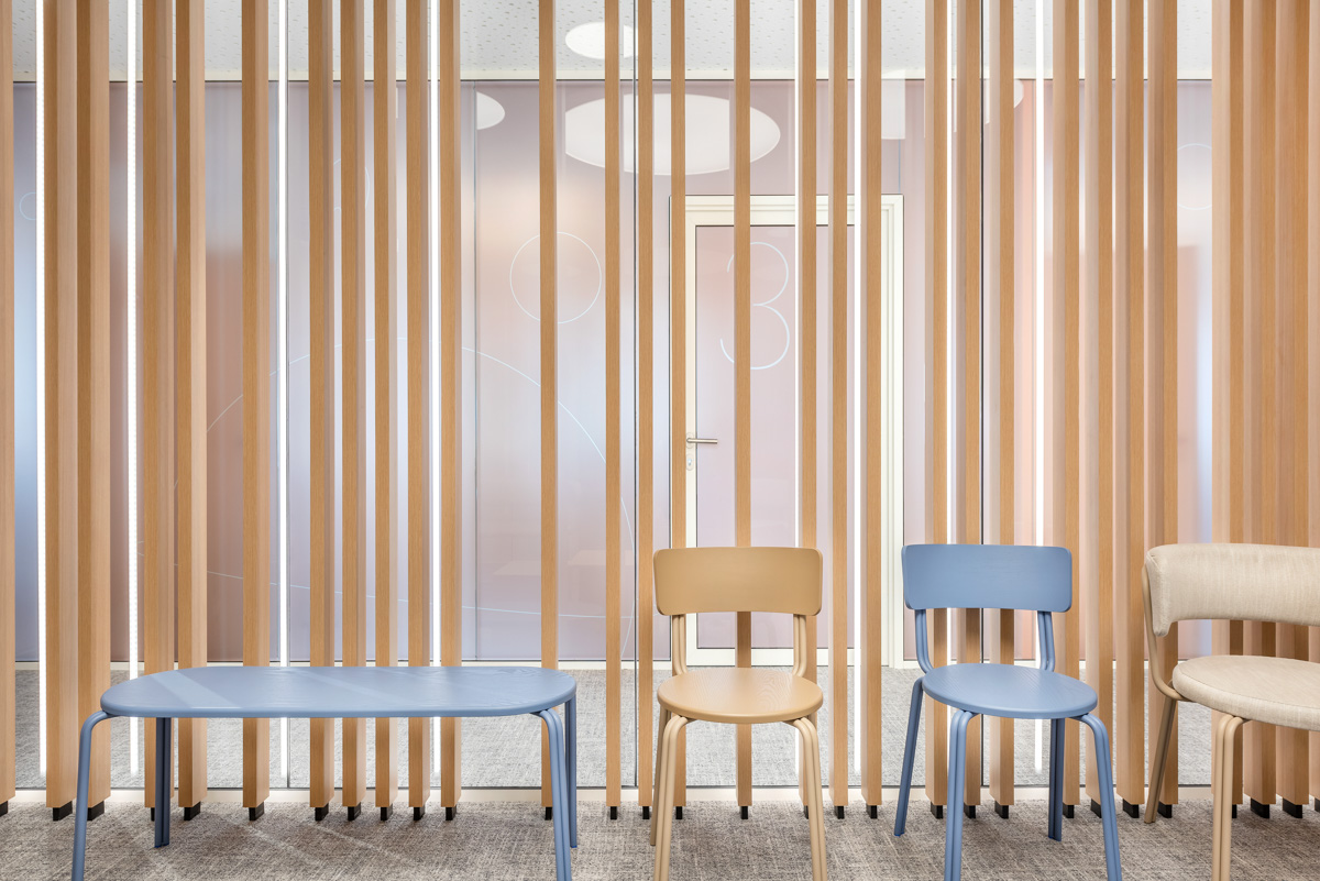 Pastellfarbige Stühle und eine Bank stehen entlang einer Glaswand mit vorgesetzten Holzlamellen