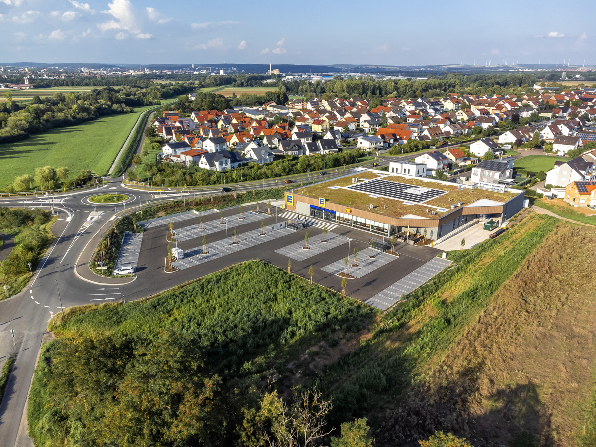 Luftbild eines Supermarktes mit Parkplätzen vor einer ländlichen Ortschaft im Sonnenuntergang